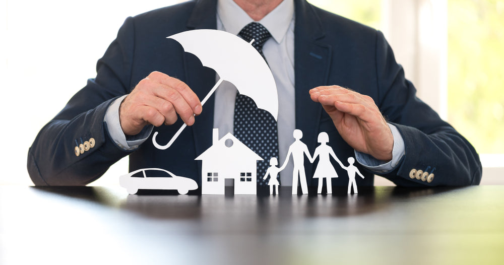 A businessman holding a paper umbrella over cutouts of a car, house, and family, symbolizing insurance and financial protection.