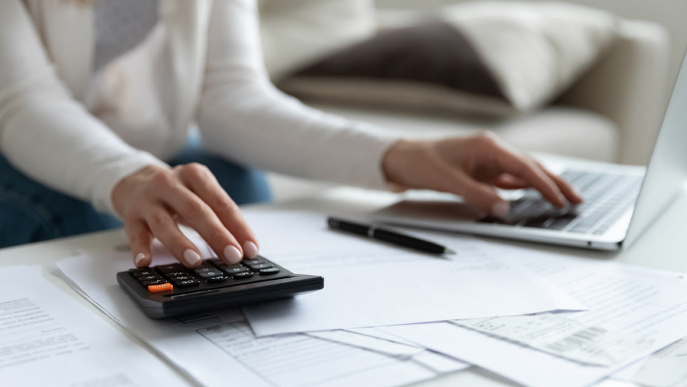 Person using a calculator and laptop on a desk with financial documents, pen, and invoices, working on budget calculations.