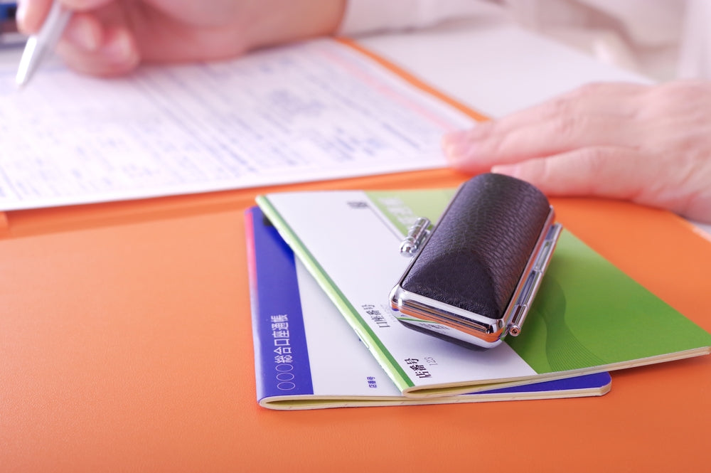 A person filling out paperwork with a pen, alongside a stack of Japanese bankbooks and a seal case on an orange folder.