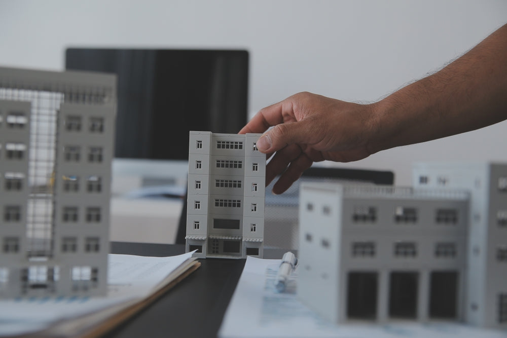 Architect's hand placing a building model on a desk with other models and architectural plans nearby.